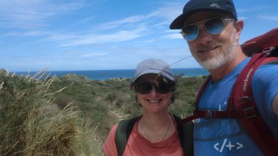 A photo of Saira and Chris with dunes and sea in the background. Chris has a grass straw in his teeth.
