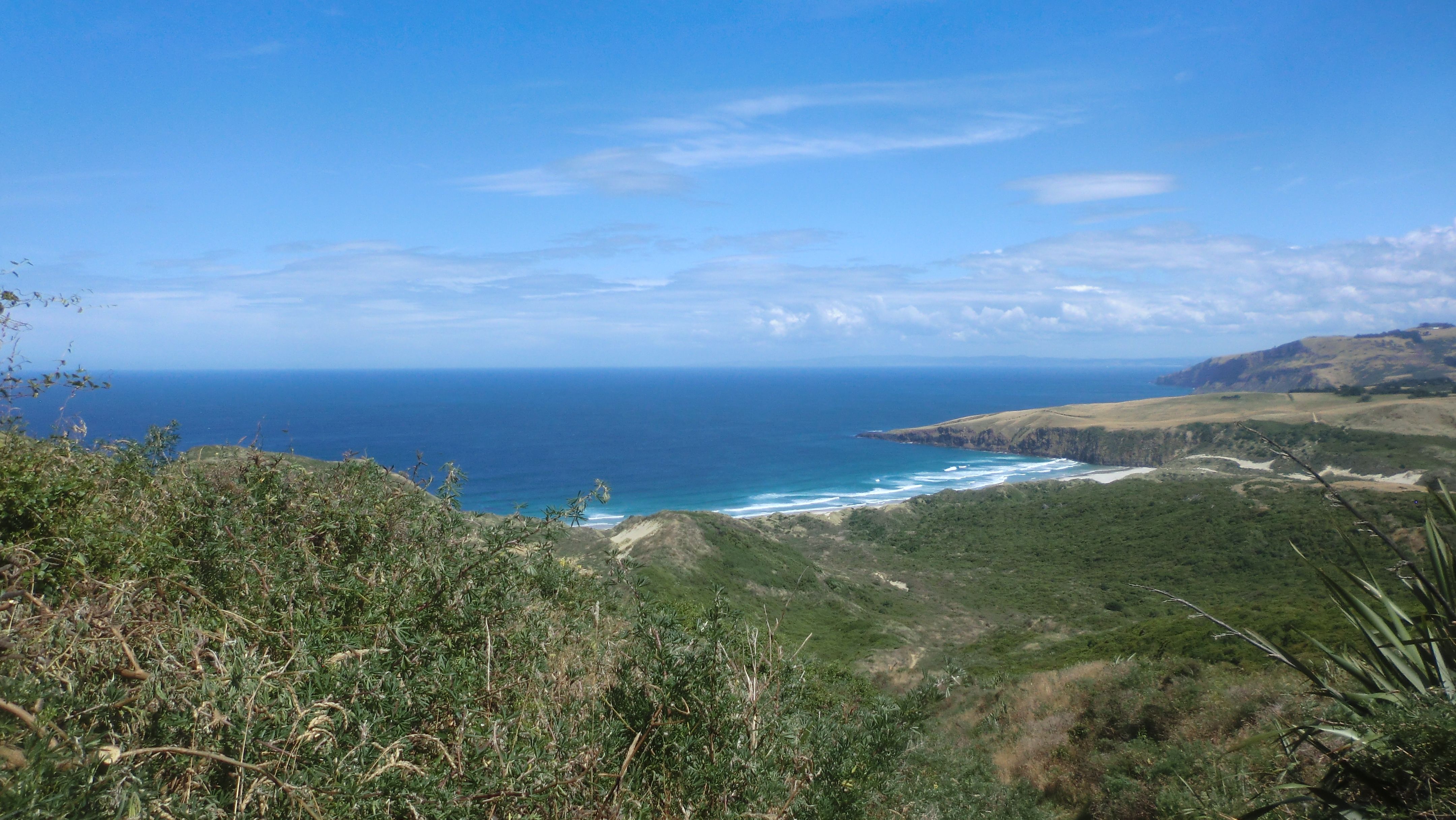 Photo - clear blue sky, low clouds in the distance, blue southern Pacific ocean, dry coastal pasture along the peninsula, dunes now visible in the distance, and a long descent to the beach.
